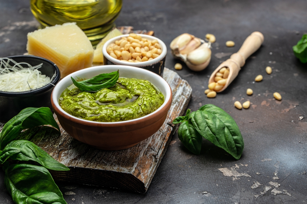 Molho pesto dentro de tigela marrom ao lado de queijos e folhas de manjericão