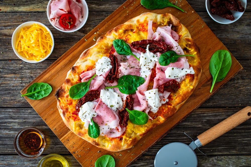 Pizza romana retangular recheada com queijo, manjericão e presunto em cima de tábua de madeira