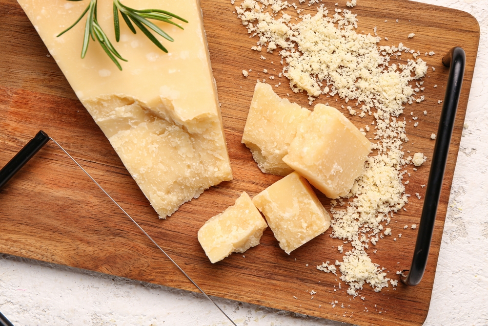 Queijo parmesão ralado e pedaços de queijo parmesão cortados em cubos em cima de tábua de madeira